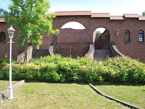 Blick auf Schlossruine - Treppen führen zur Besucherterrasse im Backsteinbau. Im Vordergrund Grünfläche für Bänke.