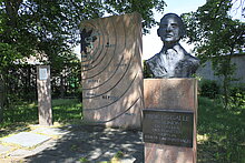 Im Vordergrunud sieht man ein Denkmal von Johann Gottfried Galle. Man sieht sein Gesicht und einen Teil seines Oberkörpers auf einer Stele mit einer Informationstafel. Im Hintergrund sieht man ein Denkmal welches unser Sonnensystem darstellt.