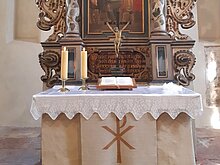 Dies ist ein Bild vom Inneren der Kirche, zu sehen ist der Altar, im Vordergrund steht ein Tisch mit Kerzen und im Hintergrund ist ein Gemälde mit feinen Verzierungen zu sehen.