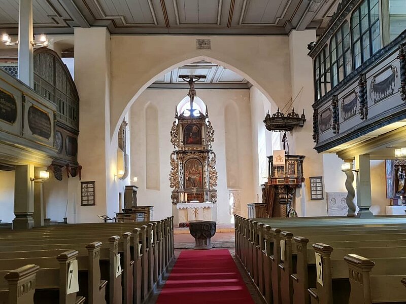 Hier sieht man den Innenraum der Kirche. Rechts und Links sind Sitzbänke zu sehen, außerdem kann man die obere Empore erkennen. Im Hintergrund sieht man den mit schönen Schnitzereien versehenen großen Altar. 