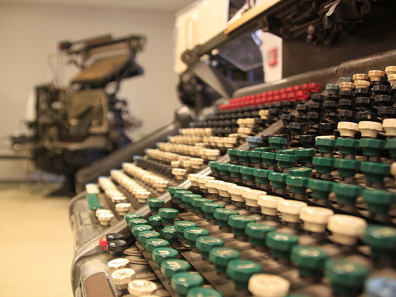 Im Vordergrund sieht man viele Tasten einer Art Schreibmaschine, dem so genannten Taster . Im Hintergrund steht eine große Maschine.
